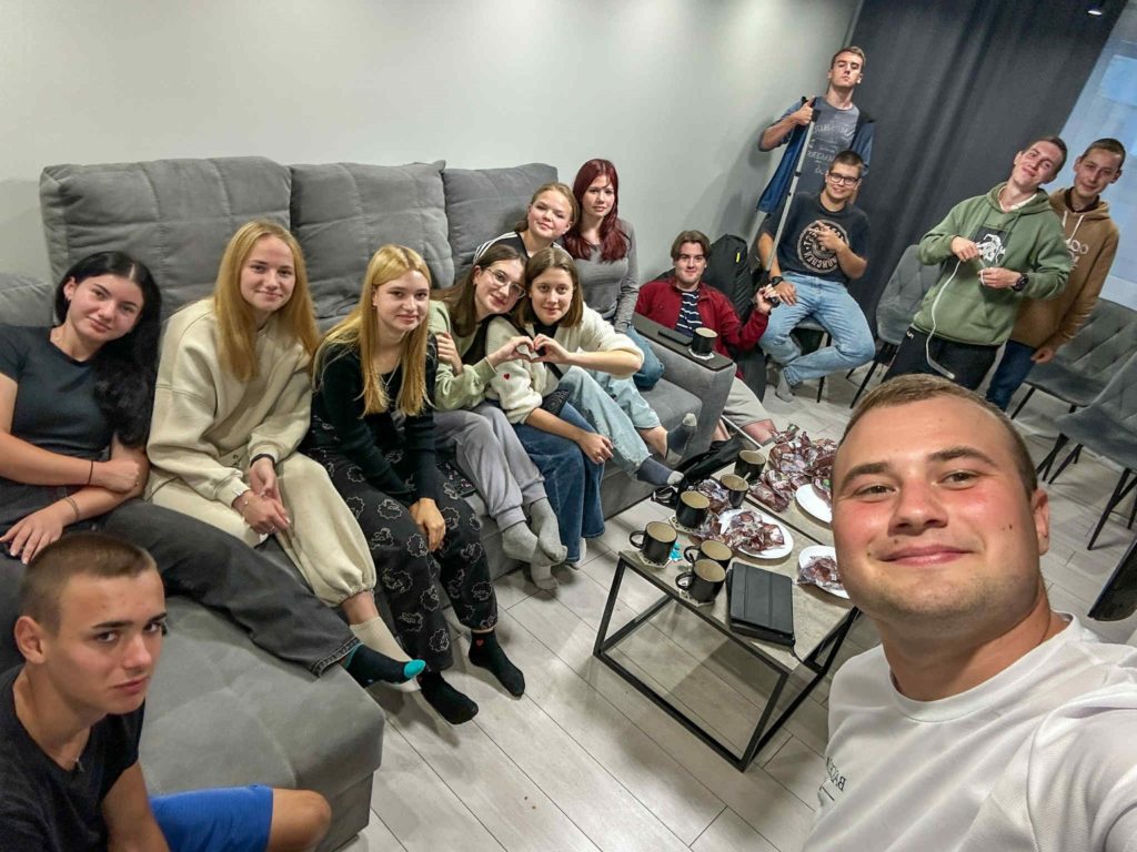 Group of young people sitting together on sofas indoors, smiling at the camera with snacks and drinks on the table.