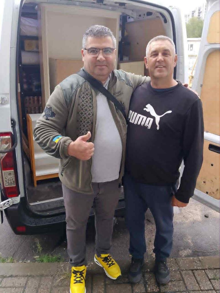 Azat and Pastor Andrew standing  side-by-side with their arms round each other at the back of a van loaded with fresh aid supplies