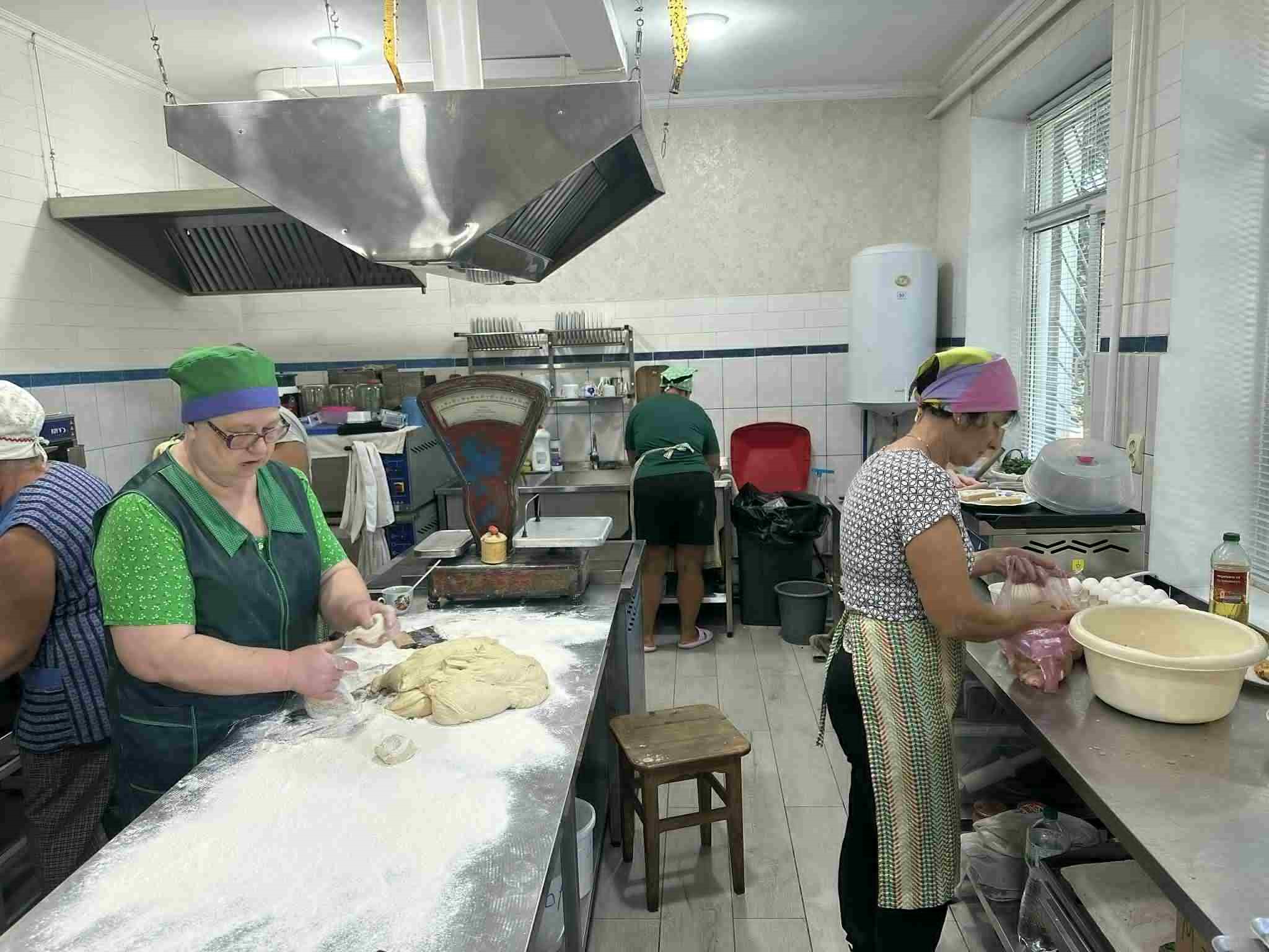 Several women working in the bakery kitchen, shaping dough and preparing ingredients on stainless steel counters.