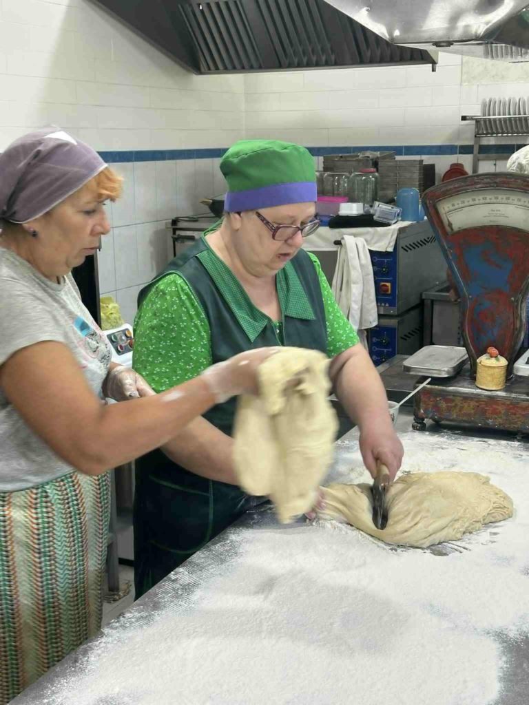 Two women in the bakery preparing dough on a floured table, one cutting the dough while the other assists.