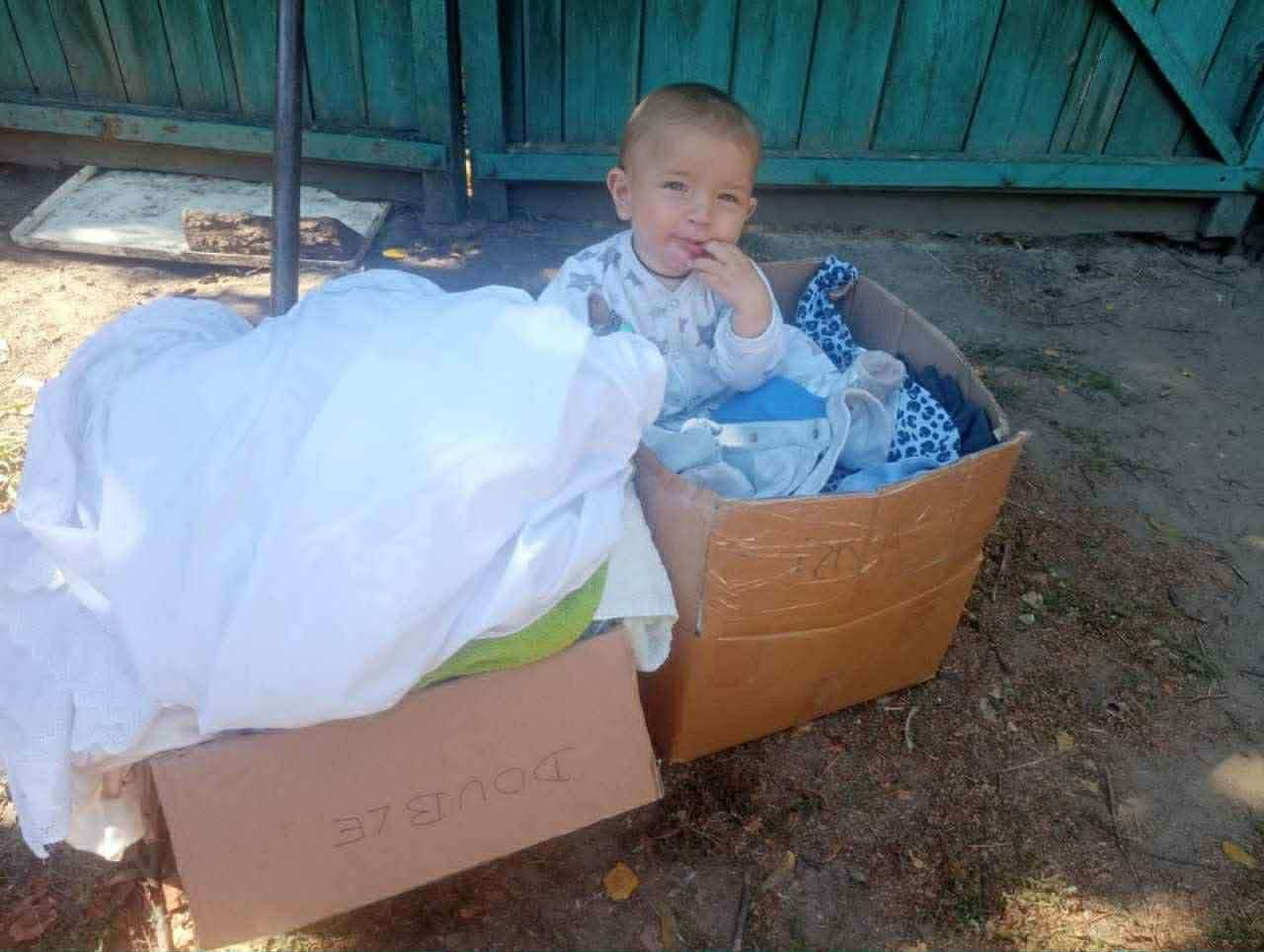 A small child sits in a cardboard box filled with donated clothes beside another box of folded bedding.