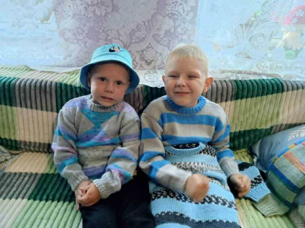 Two young boys wearing knitted sweaters sit on a sofa, smiling, with donated clothes around them.