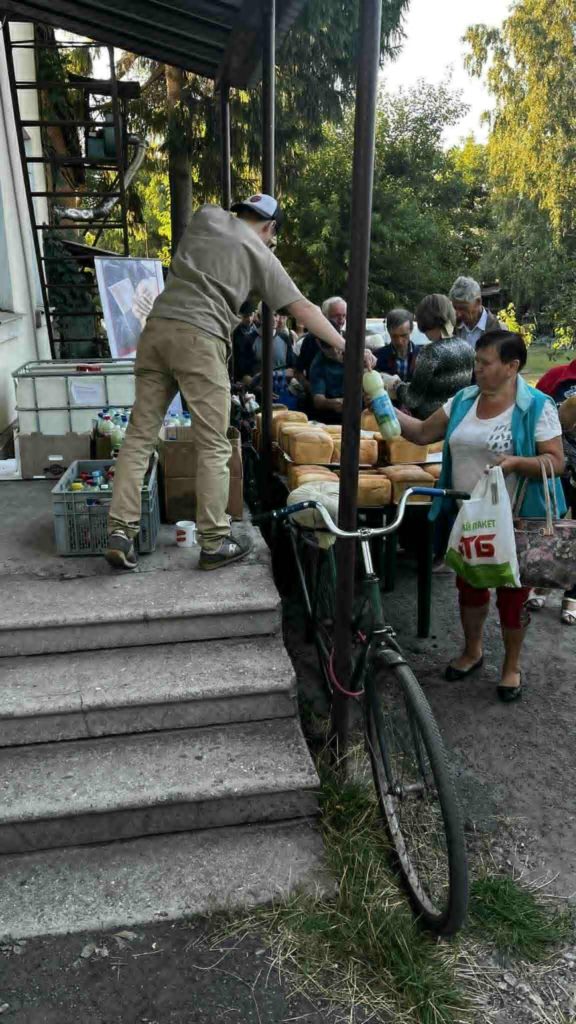 Volunteers hand out bread and bottles of shampoo to people waiting in line outside.