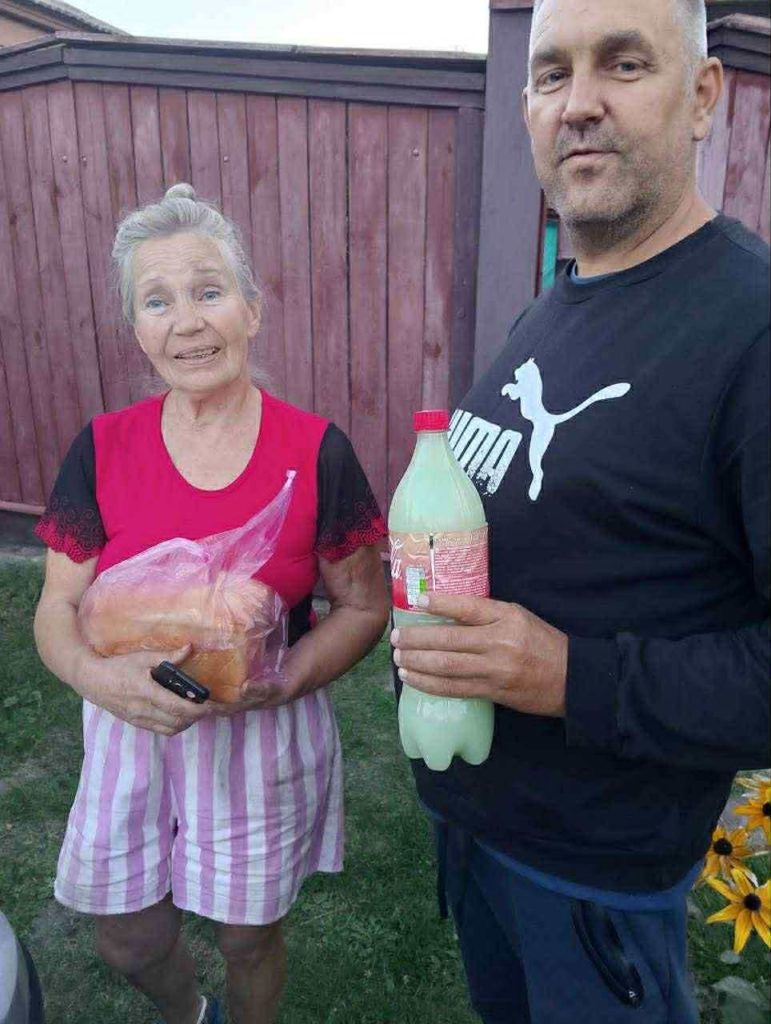 An older woman and man hold donated bread and a bottle of shampoo, smiling for the camera.