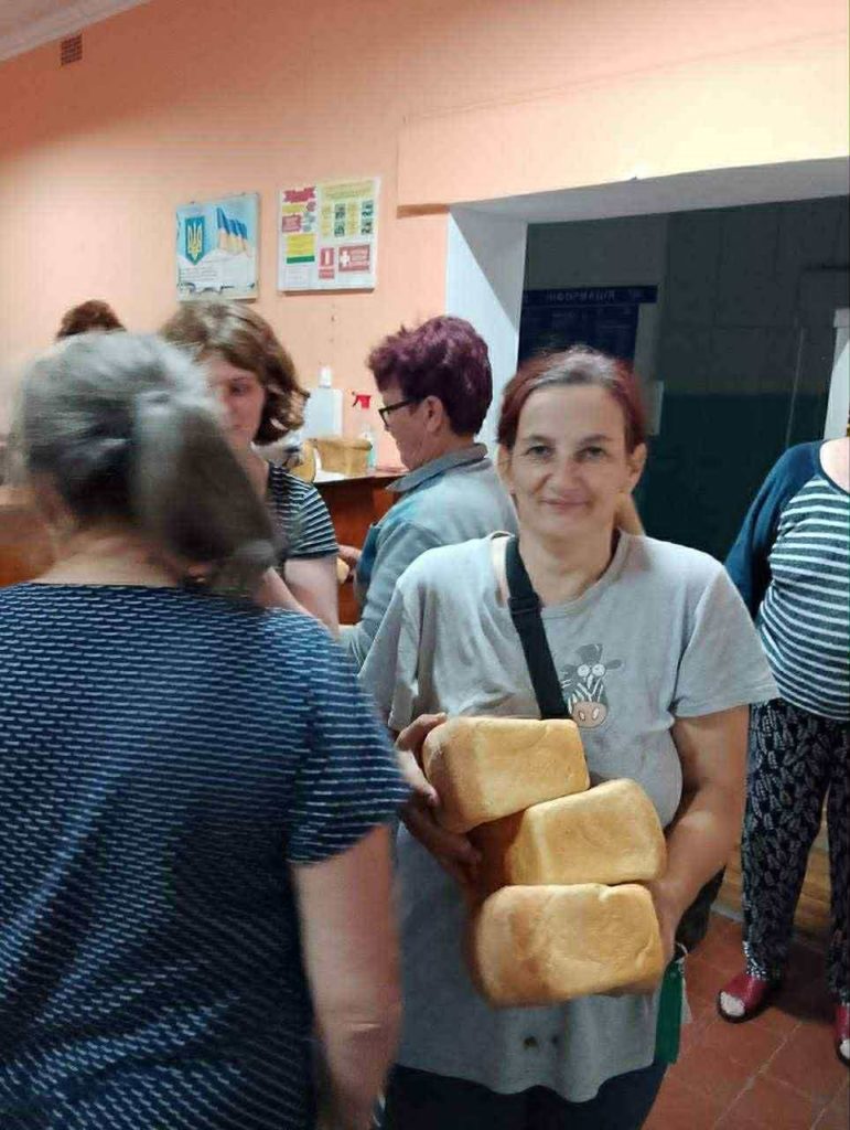 Smiling woman holding multiple loaves of bread while others queue behind her at the Centre