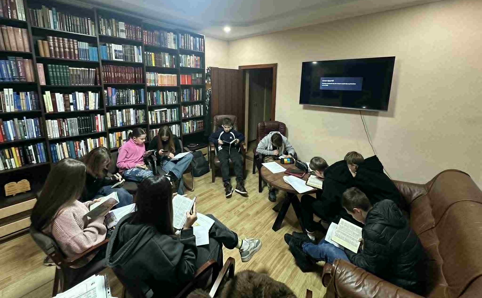 A group of teenagers sitting in a library room at a church studying the Bible together, with bookshelves lining the wall and a large TV screen at the front.
