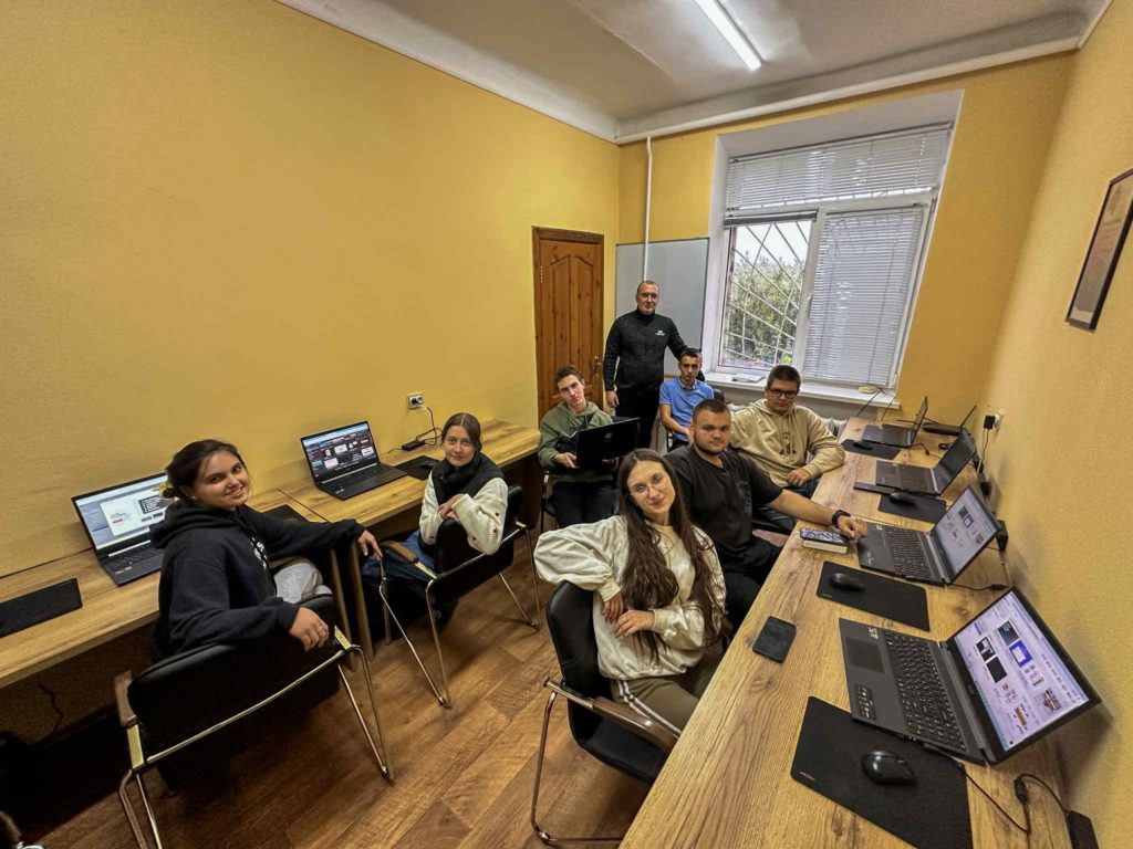 Teenagers and young adults sitting at laptops in a computer club classroom, learning digital skills