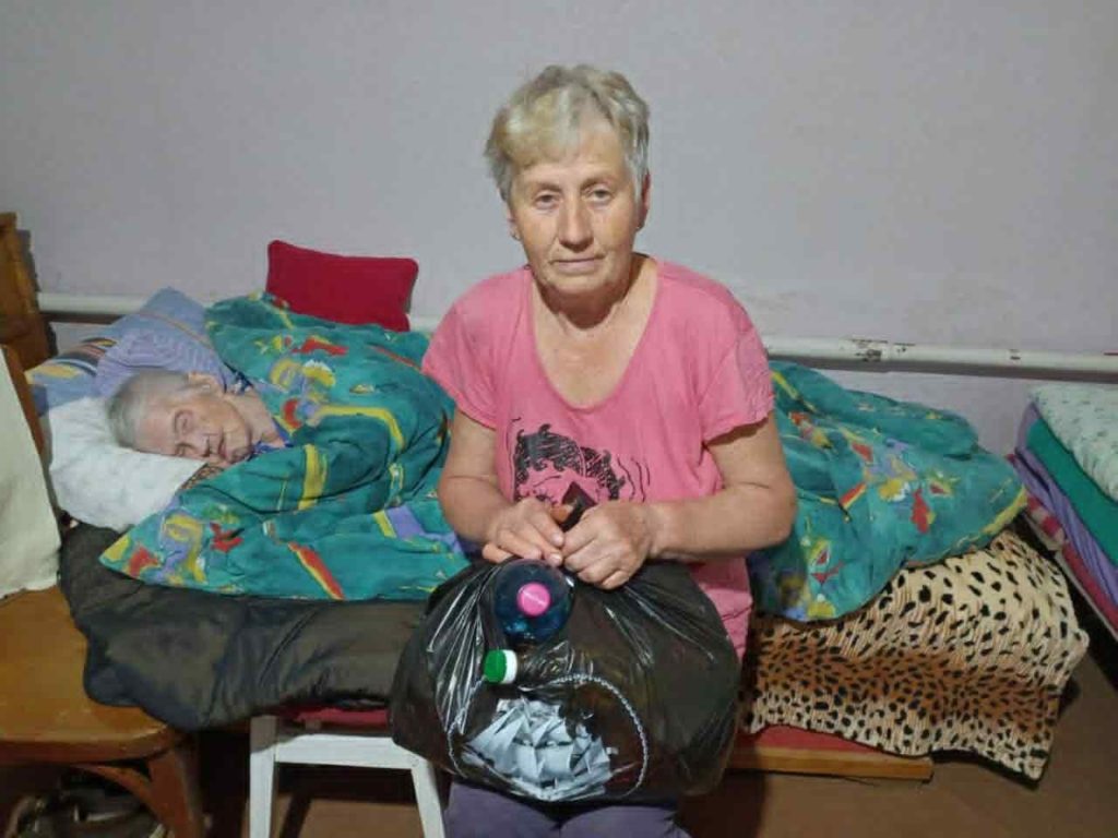 Elderly woman sitting beside a bedridden person, holding a black bag of donated food and essentials.