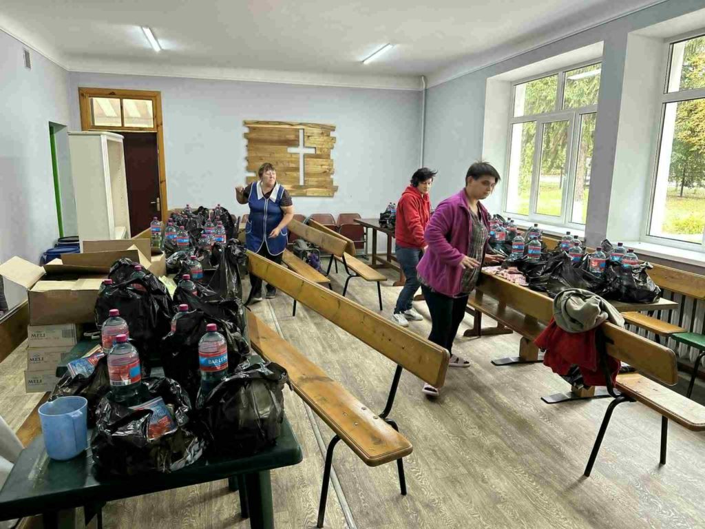 Volunteers preparing black bags filled with food and bottled water inside a large room with benches and tables.