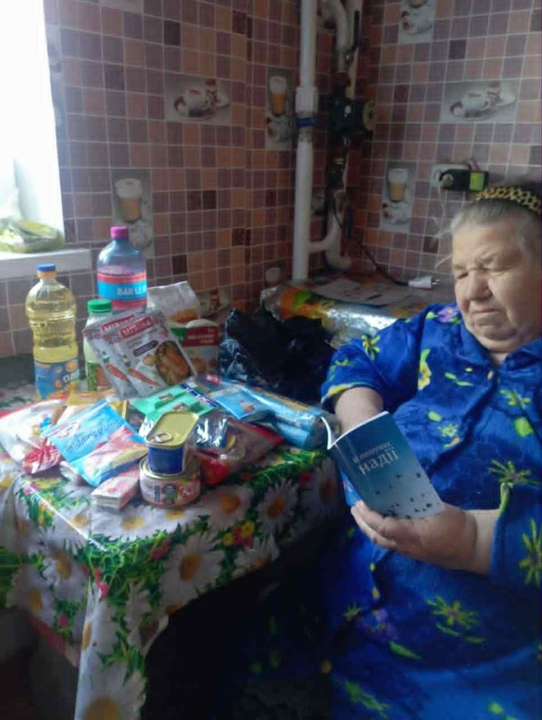Elderly woman sitting at a kitchen table with donated food items, reading a booklet titled “John’s Gospel.”