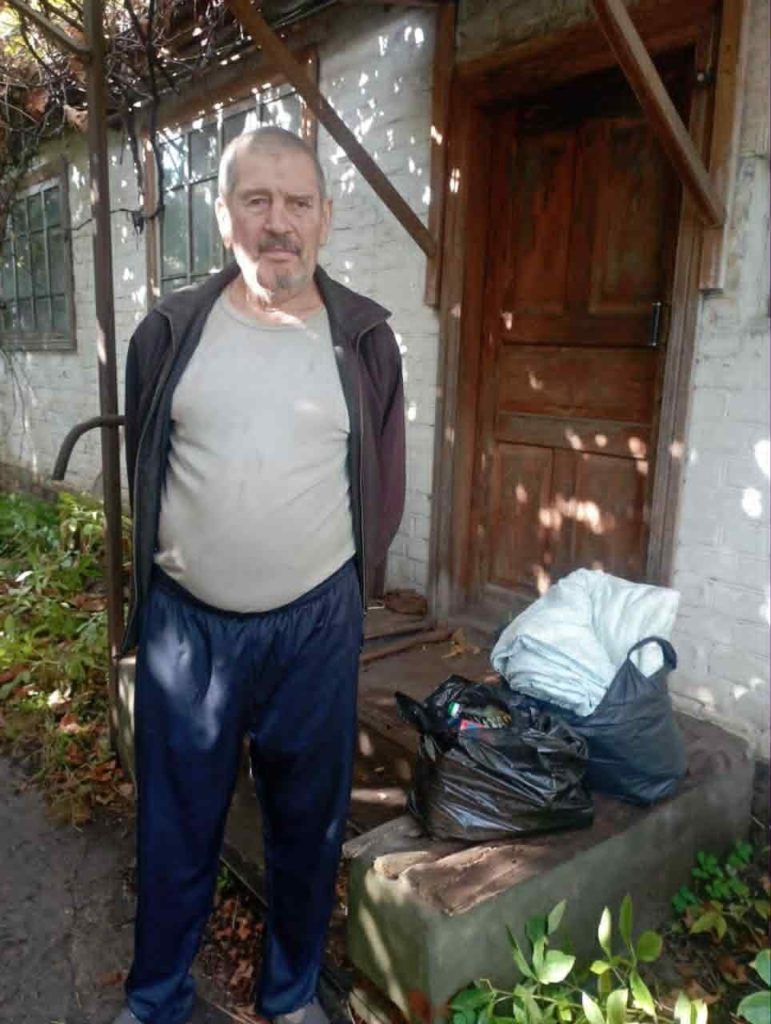 Elderly man standing outside his home next to black bags filled with donated food and blankets.