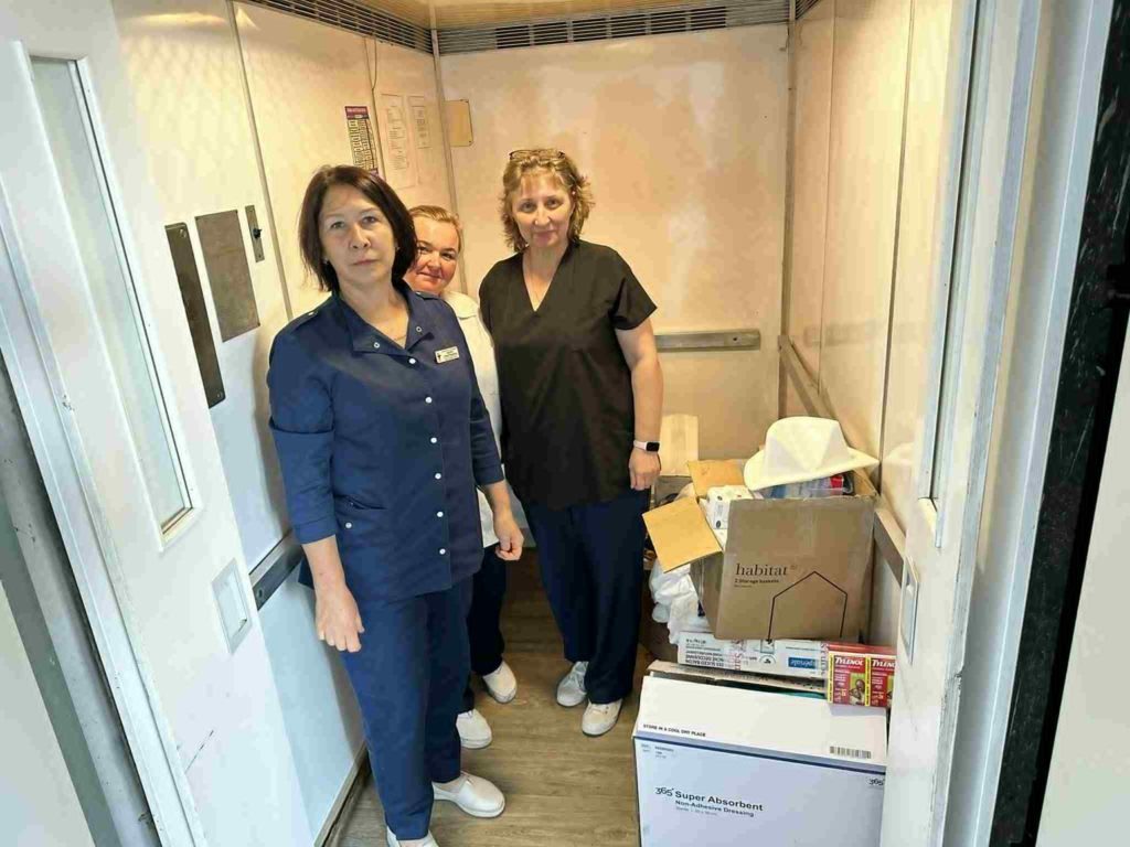 Three healthcare workers standing beside boxes of donated medical supplies inside a hospital elevator.