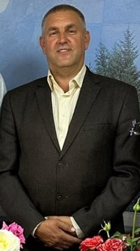 A portrait of a smiling Pastor Andrew wearing a dark suit, standing in front of a decorative floral display and a background with trees.