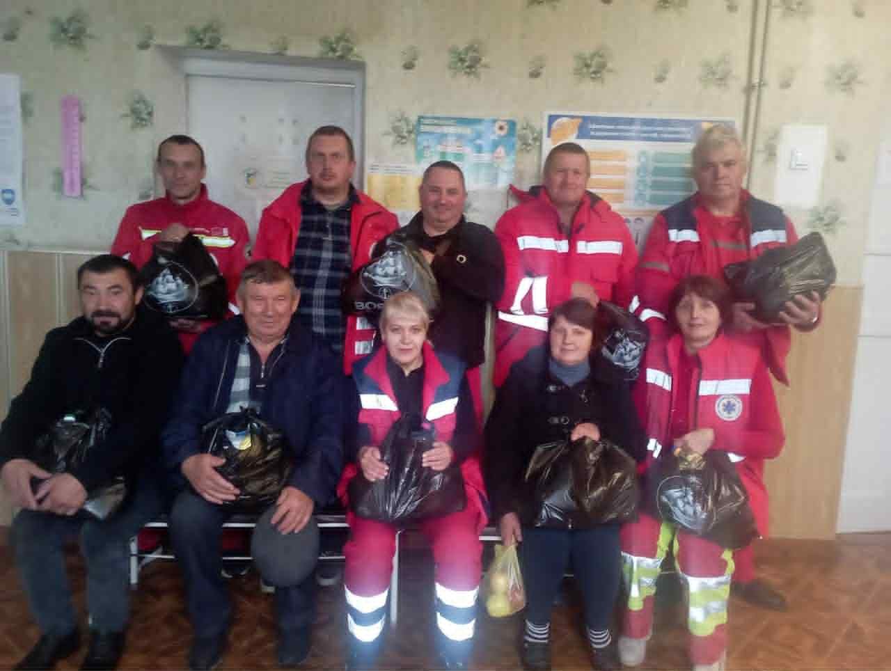 Group of emergency workers and volunteers posing indoors with black bags of donated food and essentials.