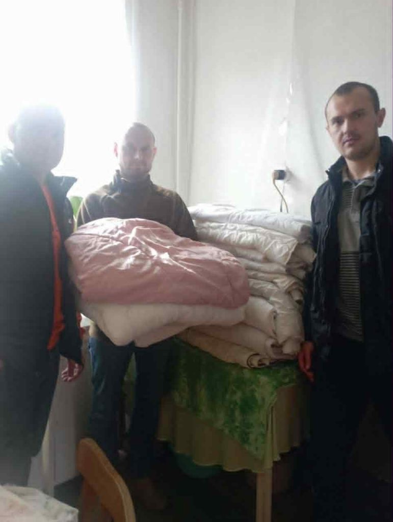 Three men standing indoors holding and stacking donated bedding and blankets for distribution to refugees.