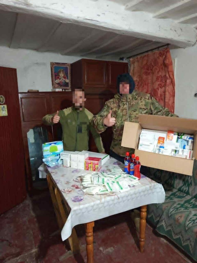 Two soldiers standing indoors with a box of donated medical supplies and hygiene products displayed on a table.