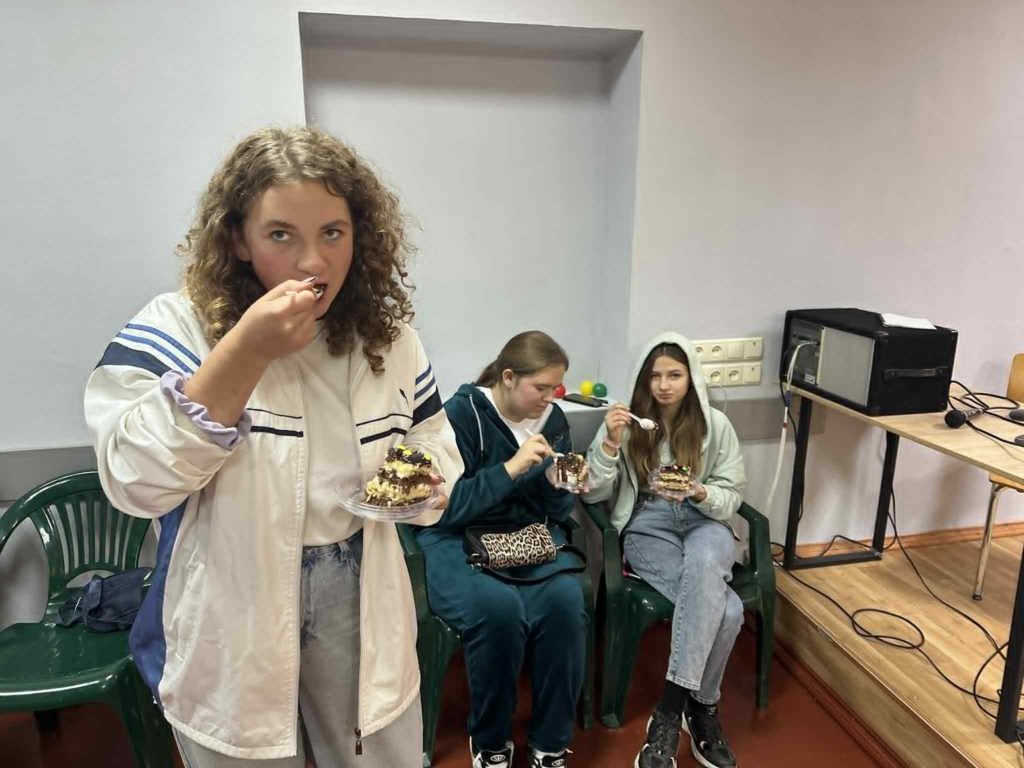 Three teenage girls eating cake while sitting and standing in a youth club room.