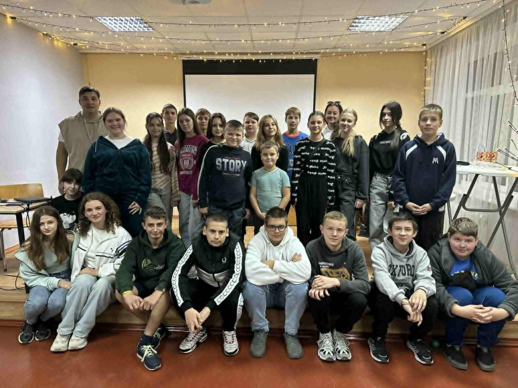Large group of teenagers and young adults posing for a group photo at a youth club, with string lights in the background.