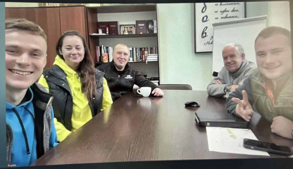 Group of five team smiling during an online team meeting with Peter, seated around a wooden conference table.