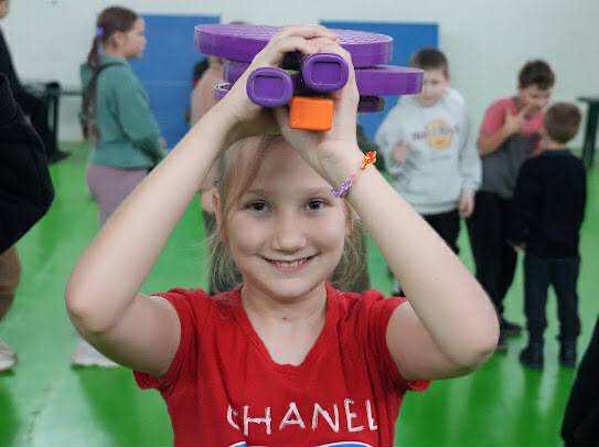 Smiling girl at a the autumn camp holding some plastic tennis racquets above her head, with other kids playing in the background.