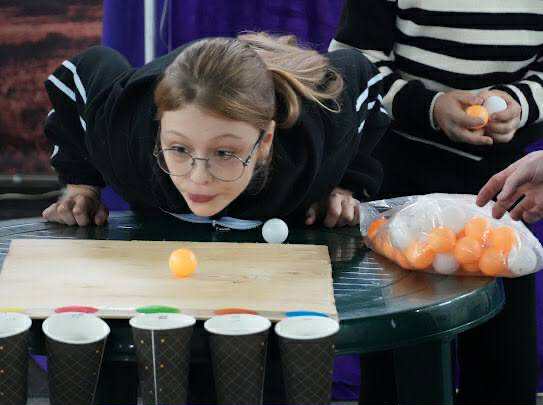 Young girl playing a fun game at the autumn camp, blowing a ping pong ball toward paper cups on a table.