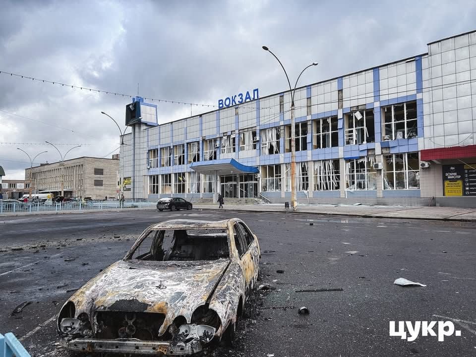 Burned-out car in front of the damaged Sumy railway station, with shattered windows and destruction from a missile strike.