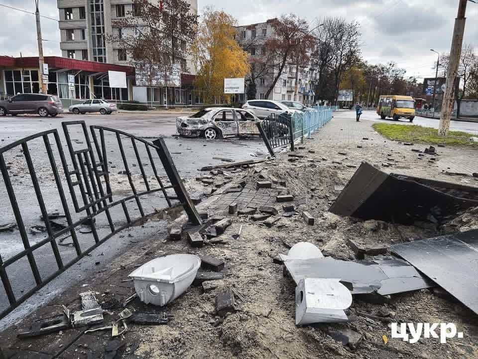 Burned-out car and shattered debris scattered across a street in Sumy after a Russian airstrike, with damaged buildings and fencing visible.
