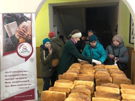 Volunteers distribute freshly baked loaves of bread to people standing in line beside a “Bread of Life” banner.