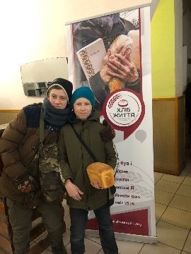 Two boys in winter clothing smile while holding a loaf of bread in front of a “Bread of Life” banner.