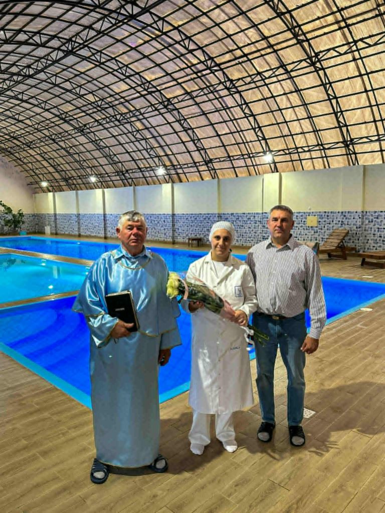 Pastor Sergey stands with two people beside an indoor pool, including a woman dressed in white holding flowers before her baptism.