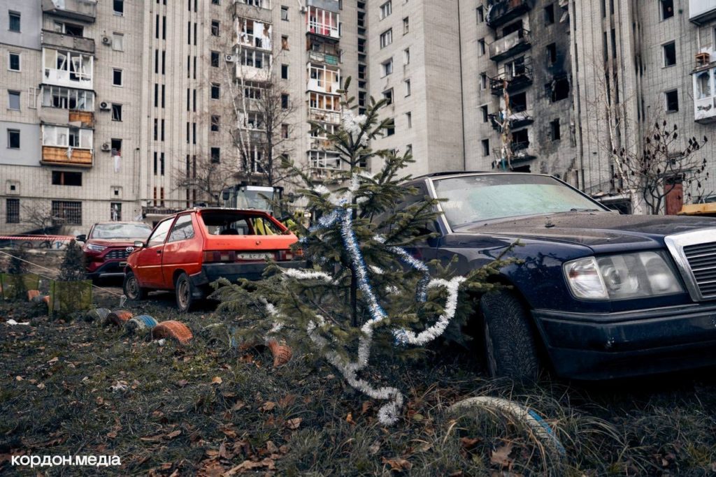 Burned and damaged cars outside a shelled apartment block in Okhtyrka, with a small decorated Christmas tree in front.
