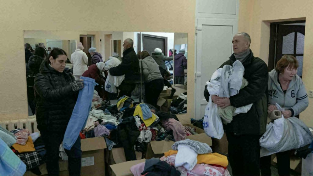 People sorting through donated clothing and blankets in a room filled with boxes and tables of items