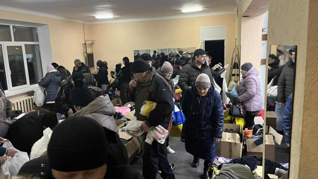 A crowded donation room where people are selecting warm clothing and blankets from boxes and tables.