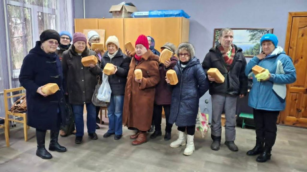 Group of elderly men and women standing indoors, each holding a loaf of donated bread.