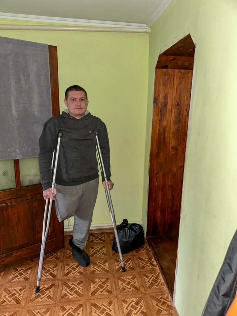 Man with one leg standing on crutches inside his home next to a bag of donated aid.