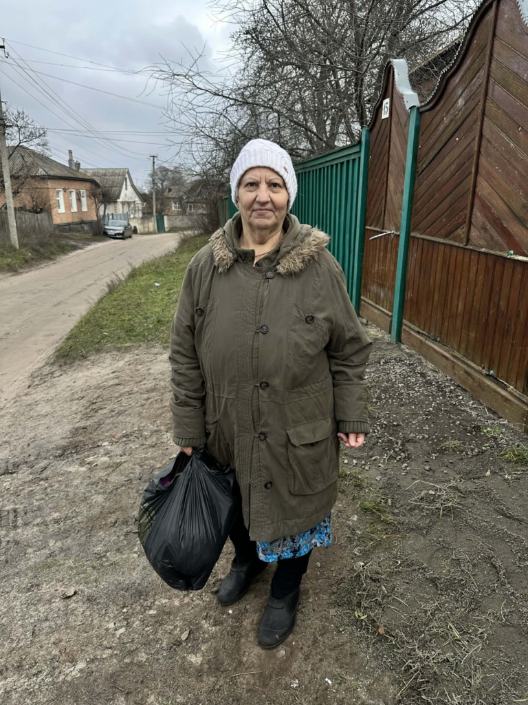 Elderly woman standing on a village roadside holding a black bag of humanitarian aid.