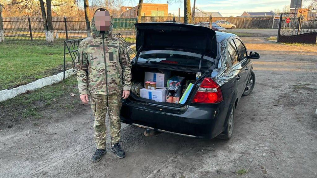 Soldier in camouflage standing next to an open car boot filled with food supplies and humanitarian items.