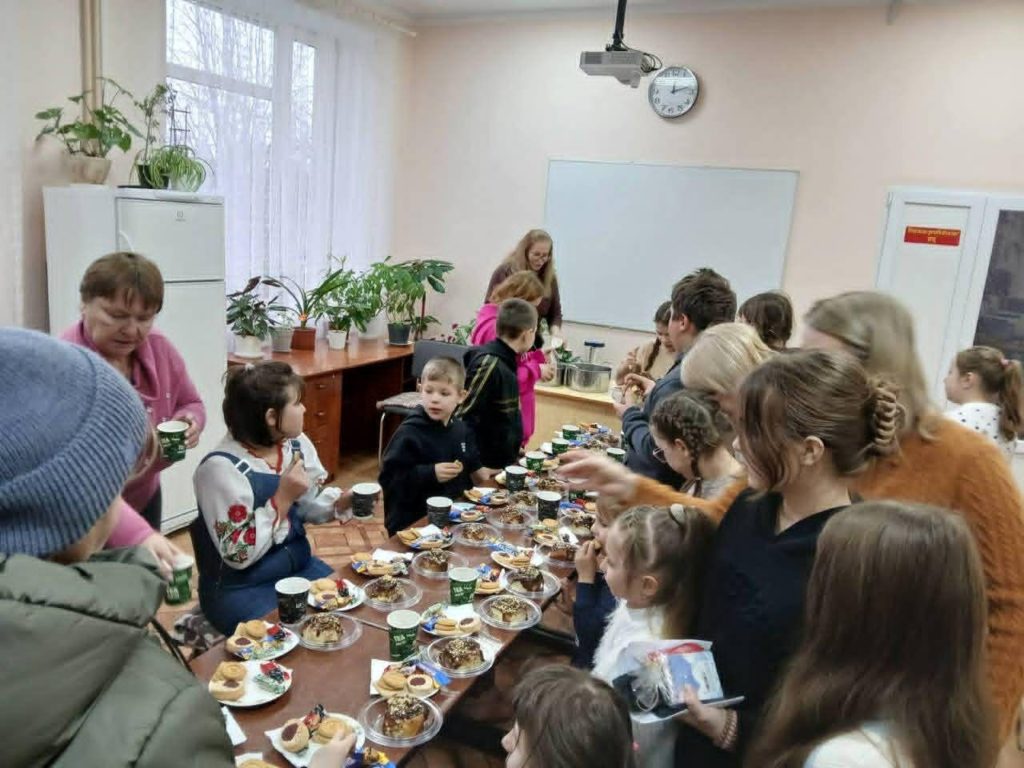 Children and adults gathered around a long table indoors, sharing tea and snacks together.