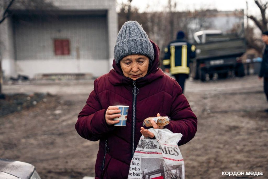 Elderly woman in a maroon coat and grey knitted hat holding a cup of hot drink and packaged snacks in a devastated outdoor area.