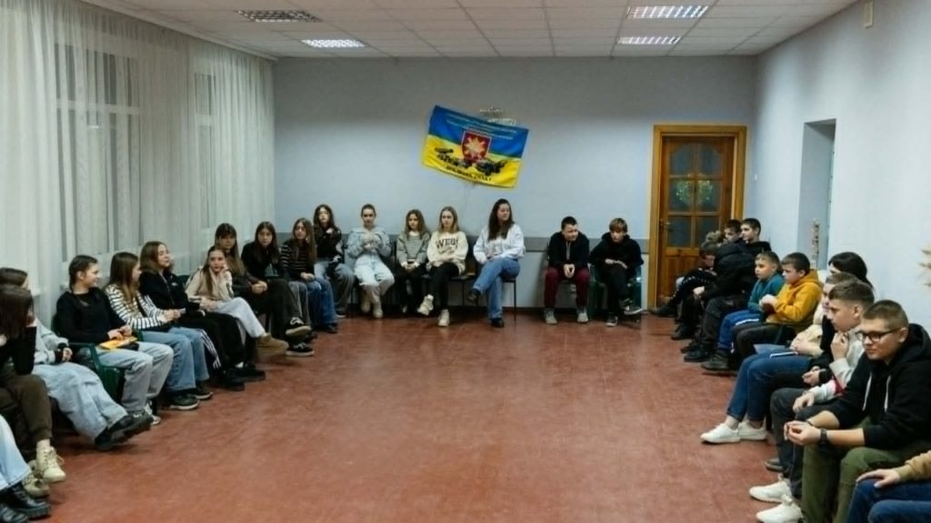 Teenagers seated in a wide circle around the edges of a room at the Centre during a youth meeting.
