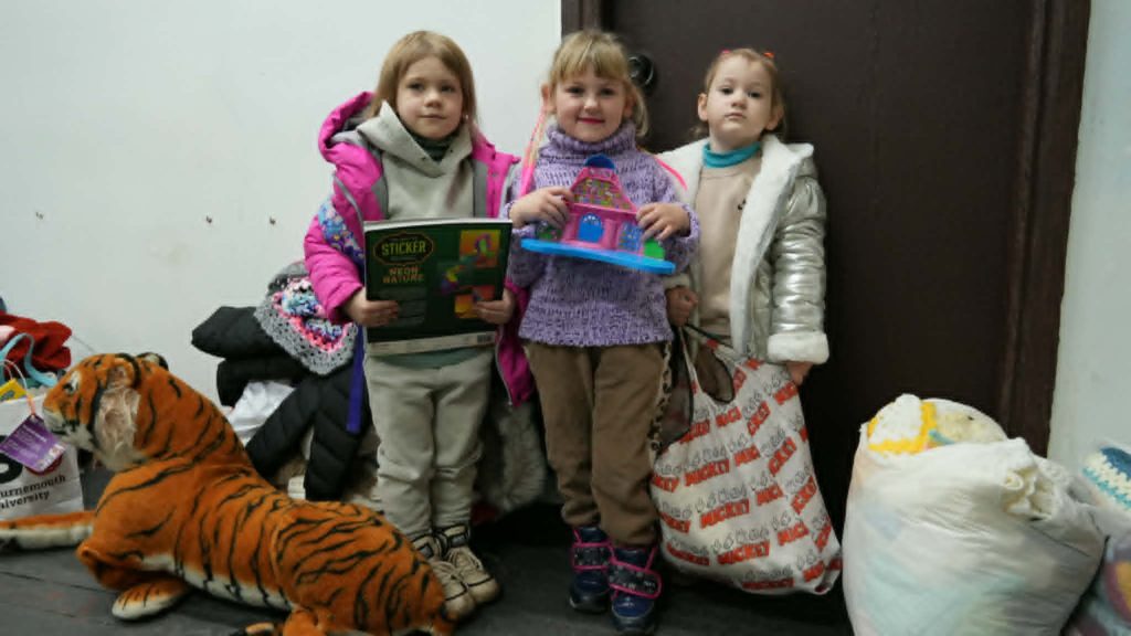 Three young girls standing indoors holding donated toys and books, surrounded by bags and soft toys.