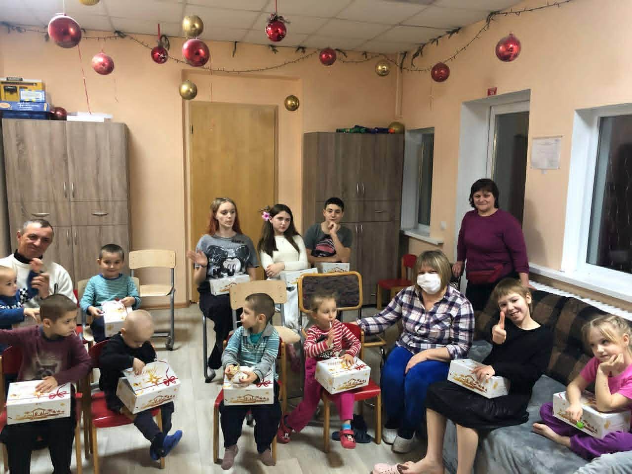 Children and carers seated indoors holding Christmas gift boxes during a festive gathering.