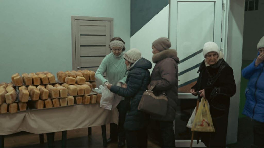Women distributing loaves of bread to elderly people indoors.