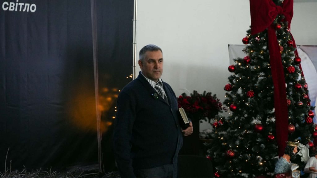 Pastor Andrew holding a Bible stands beside a decorated Christmas tree during a church Christmas service.