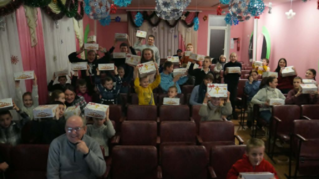 Children seated in a hall holding Christmas gift boxes during a festive event.