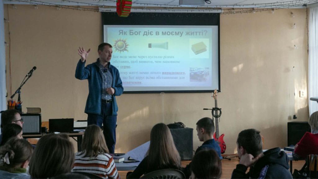 Presenter gesturing while teaching from a projected slide titled “How God acts in my life” to a seated audience.