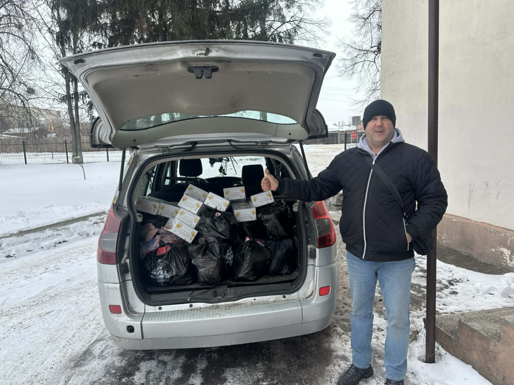 Man standing beside a car with the boot open, filled with black aid bags and food boxes, giving a thumbs-up in snowy weather.