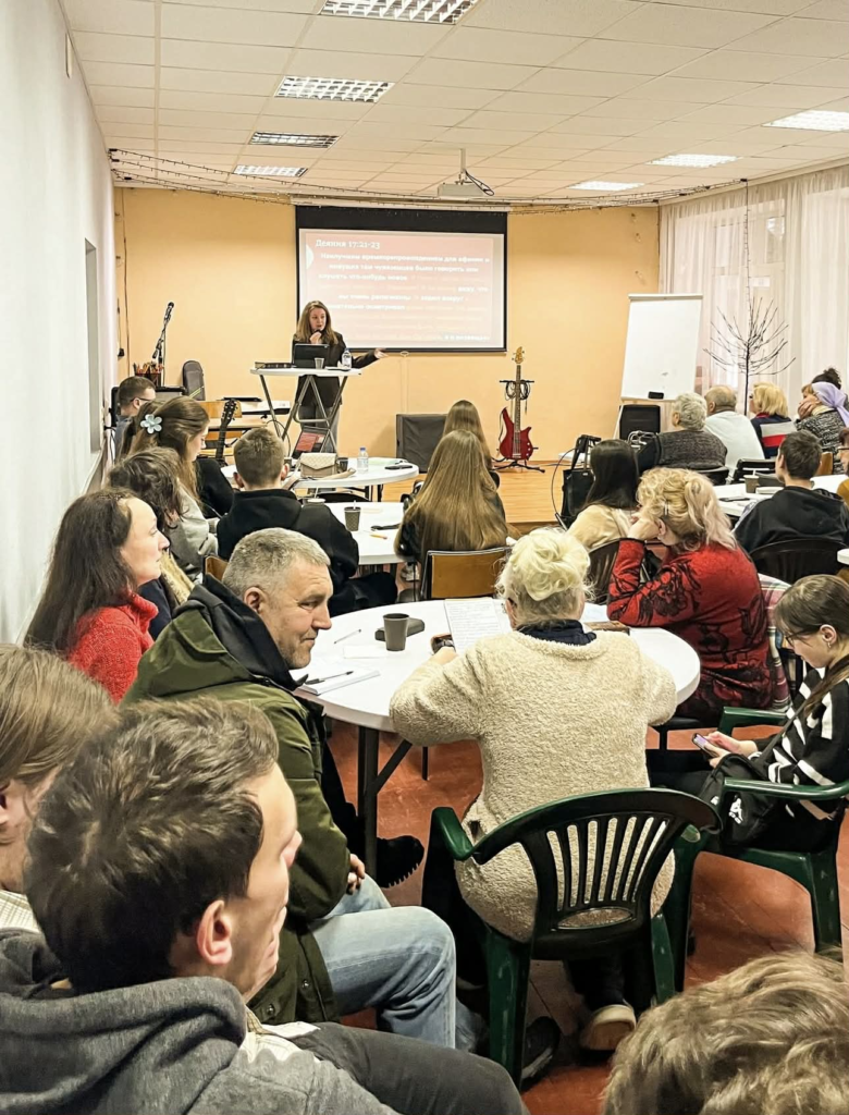 Community members attend a conference session, listening to a speaker presenting practical support and encouragement on effective evangelism during times of war.