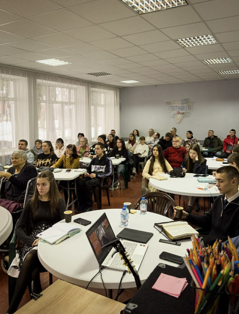 Men and women of different ages gather at tables for a conferernce workshop, learning and taking notes together in a supportive community setting.