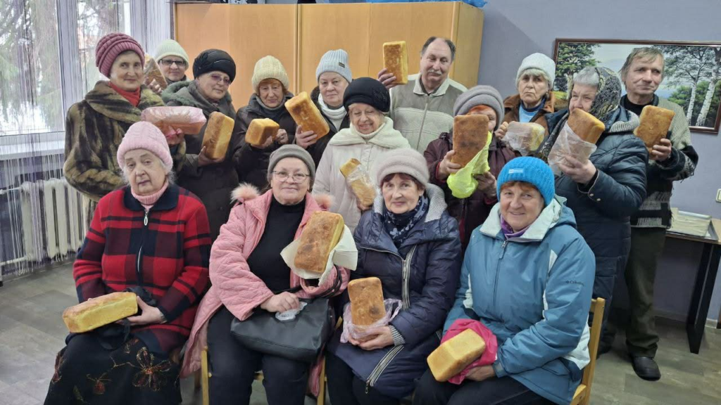 A group of elderly men and women indoors, each holding a loaf of bread received from the Centre.